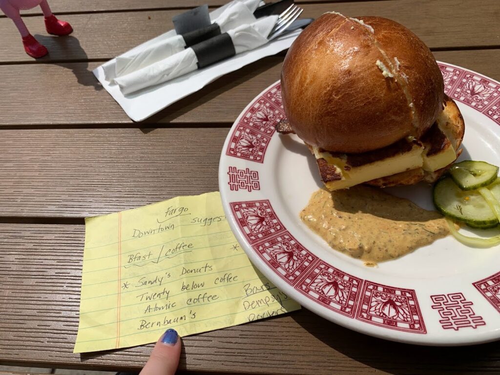 Photo of a bacon, egg, and cheese sandwich on a plate with garnish on a picnic table with a folded list next to it.