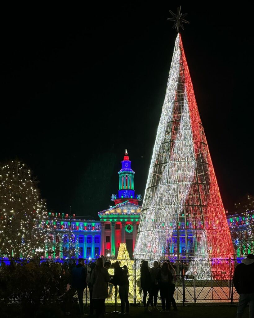 Photo of the Mile High Tree lit up with a candy cane pattern in front of a multi-colored City Council Building.